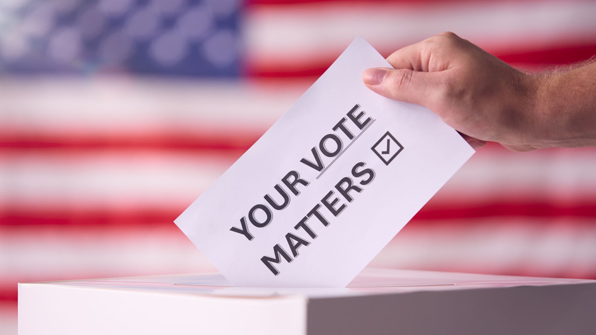 Hand Placing Ballot in Box With American Flag in Background